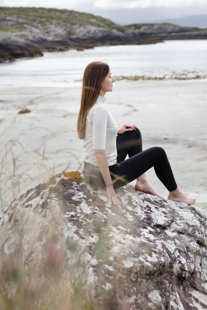 woman wearing white top and black leggings sitting on a rock on a beach