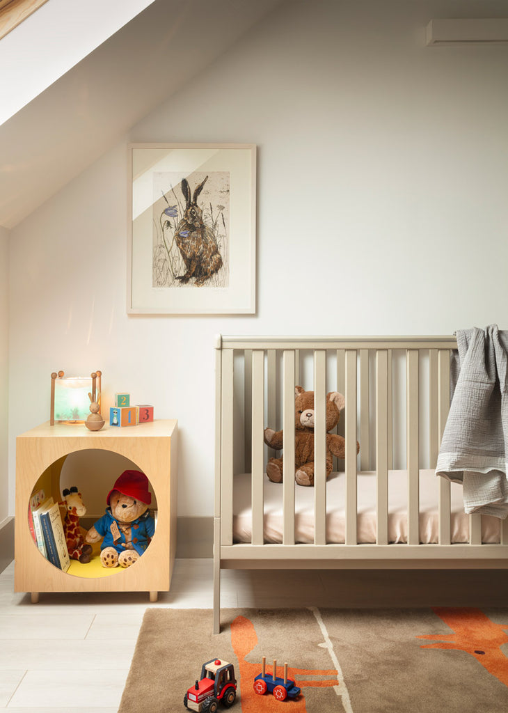 Childs bedroom with a toy shelf with a few colourful toys beside a cot with a teddy bear inside. An image of a rabbit hangs above them on the wall. 