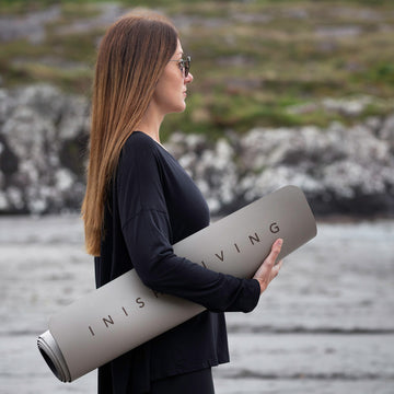 Woman stands on the beach with a rolled up high grip Inish Living exercise mat