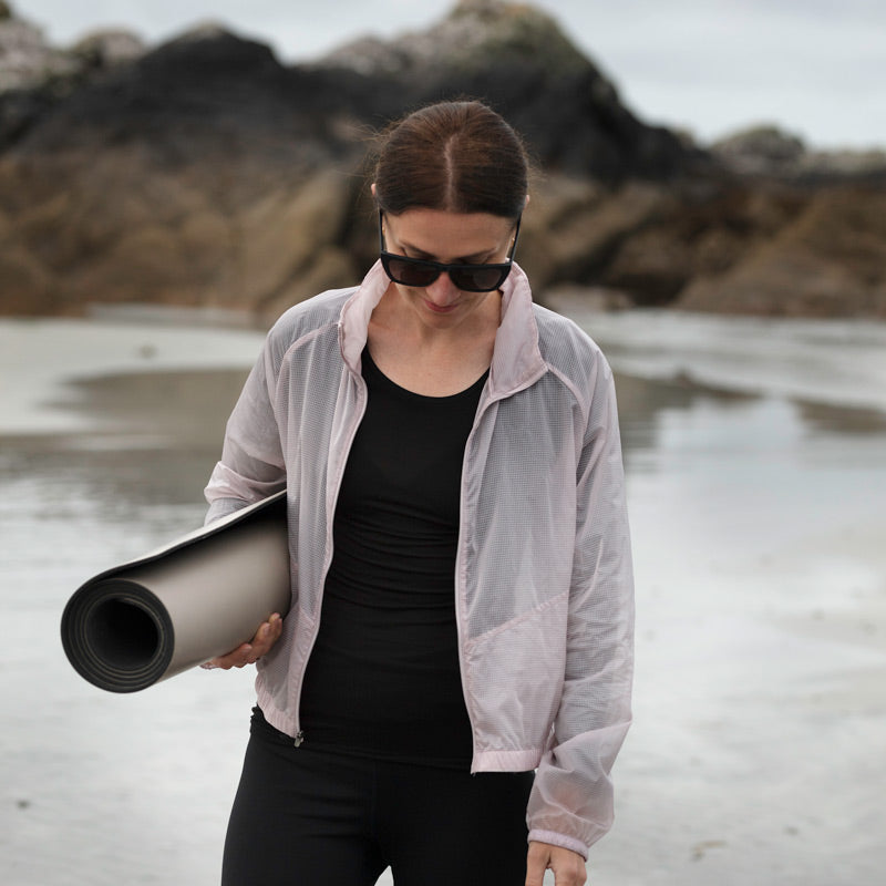 Woman stands on the beach with head down and holding a rolled up high grip Inish Living exercise mat